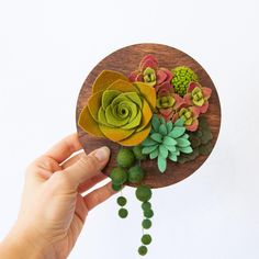 a hand holding a wooden brooch with succulents and flowers on it