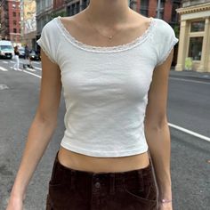 a woman standing on the side of a road with her hands in her pockets and wearing a white shirt