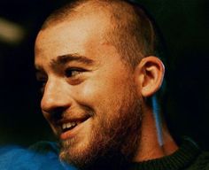 a man with a goatee smiles at the camera while wearing a blue shirt and black sweater