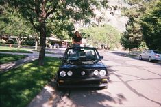 a woman standing on the hood of a parked car in front of a tree lined street