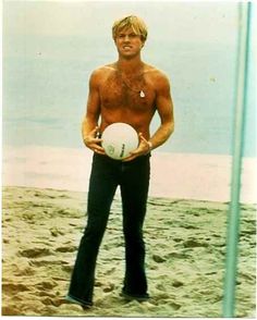 a man with no shirt holding a white ball in his right hand and standing on the beach