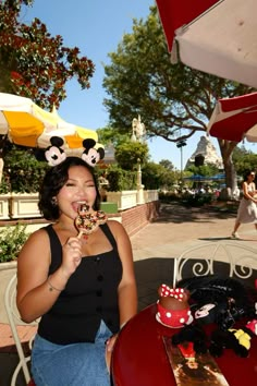 a woman sitting at an outdoor table with mickey mouse ears on her head eating cake