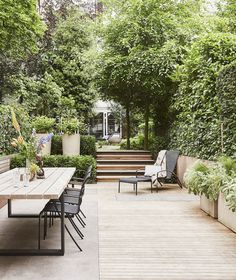 an outdoor dining table and chairs on a wooden deck surrounded by greenery with stairs leading up to the house