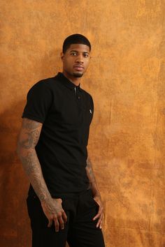 a man with tattoos standing in front of a brown wall wearing black pants and a polo shirt