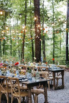 an outdoor dining table set up in the woods with hanging lights and flowers on it