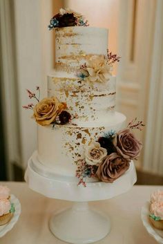 a three tiered cake with flowers on it sitting on a table next to cupcakes