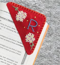 a bookmark with embroidered letters and flowers on top of an open book in the shape of a triangle