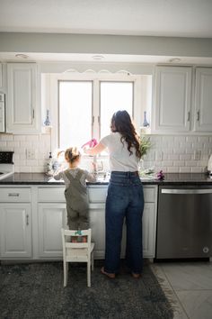 a woman standing in front of a kitchen window next to a child on a chair