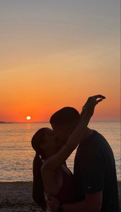 a man and woman kissing on the beach at sunset, with the sun setting in the background