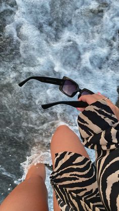 a woman sitting on top of a beach next to the ocean holding onto her sunglasses