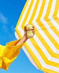 a woman holding up a can of mustard in front of an open yellow and white striped umbrella