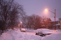 a snowy street with cars parked on the side and one car covered in snow at night