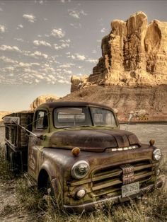 an old pick up truck sitting in the grass near some rocks and water with a mountain in the background