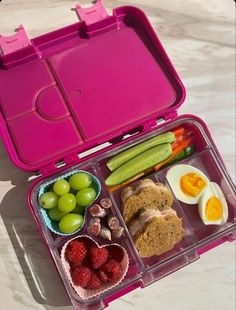 a pink lunch box filled with food on top of a white table next to an egg, cucumber, and fruit