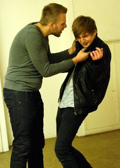 two men standing next to each other in front of a white wall and one is adjusting the jacket