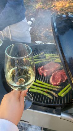 a person holding a wine glass in front of a grill with steak and asparagus on it