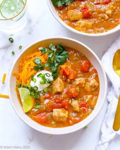 two bowls of chicken and vegetable soup on a white table with gold spoons, lime wedges and cilantro