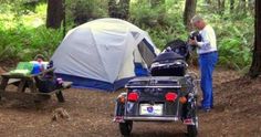 a man standing next to a motorcycle near a tent and picnic table in the woods