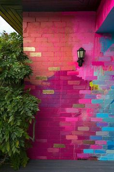 a pink and blue painted brick wall next to a green bush with a lamp on it