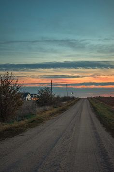 an empty dirt road in the middle of a rural area at sunset with houses on either side