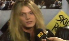 a man with long blonde hair holding a microphone and talking to the camera while wearing a black leather jacket