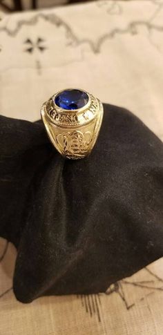 a gold and blue ring sitting on top of a black cloth covered pillow in front of a bed