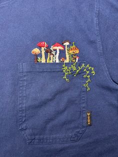 a blue shirt with mushrooms embroidered on the front pocket and in the back pocket is a green plant growing out of it