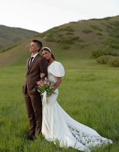 a bride and groom pose for a photo in the grass on their wedding day with mountains in the background