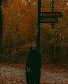 a woman standing next to a street sign in the middle of an autumn forest with leaves on the ground