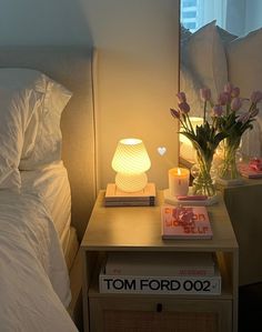 a bedside table with flowers and books on it next to a lamp that is turned on