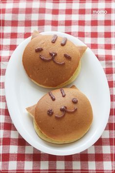 two pancakes with cats on them are sitting on a white plate next to a red and white checkered tablecloth