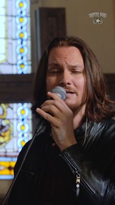 a man with long hair holding a microphone in front of stained glass windows and wearing a leather jacket