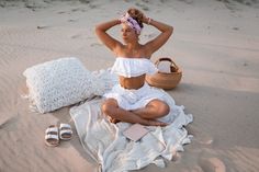 a woman sitting on top of a sandy beach next to a basket and book in her hand