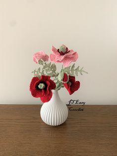 a white vase filled with red flowers on top of a wooden table next to a wall