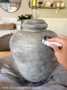 a person is painting the bottom of a large gray vase on a table with a mirror in the background