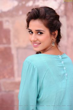 a beautiful young woman standing in front of a brick wall wearing a green blouse and gold earrings