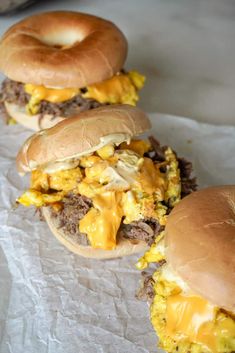 three hamburgers with cheese on them sitting on top of wax paper next to a bagel