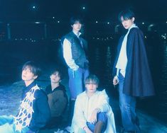 group of young men sitting on the ground in front of water at night with one man wearing a cape