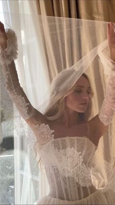 a woman wearing a wedding dress and veil in front of a window with sheer curtains