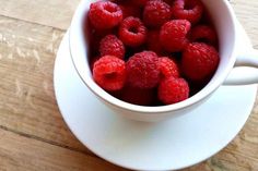 some raspberries are in a white cup on a saucer and wooden table