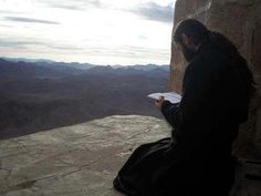 a man sitting on top of a stone wall next to a tall rock structure with mountains in the background