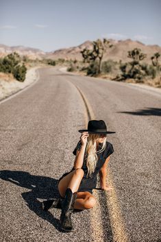 a woman sitting on the side of a road wearing a cowboy hat and black dress