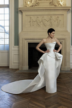 a woman standing in front of a fireplace wearing a wedding dress with an asymmetrical train