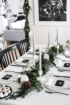 the table is set for christmas dinner with white plates and silverware, black napkins, pine cones, evergreen garland and candles