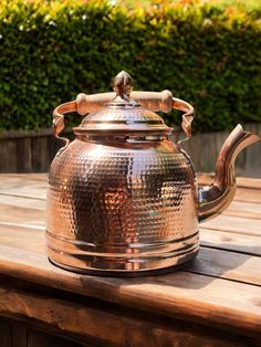 a metal teapot sitting on top of a wooden table next to a bushy area