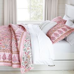 a white bed topped with pink and green comforter next to a window filled with curtains