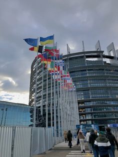 several flags are flying in the wind near a building with many windows and people walking around