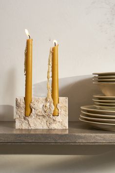 two yellow candles sitting on top of a table next to plates and bowls in front of a white wall