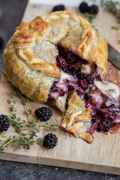 a blackberry pie is cut in half on a cutting board with berries and herbs around it