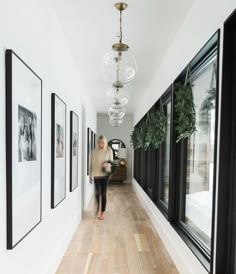 a woman walking down a long hallway with pictures hanging on the wall and plants in vases